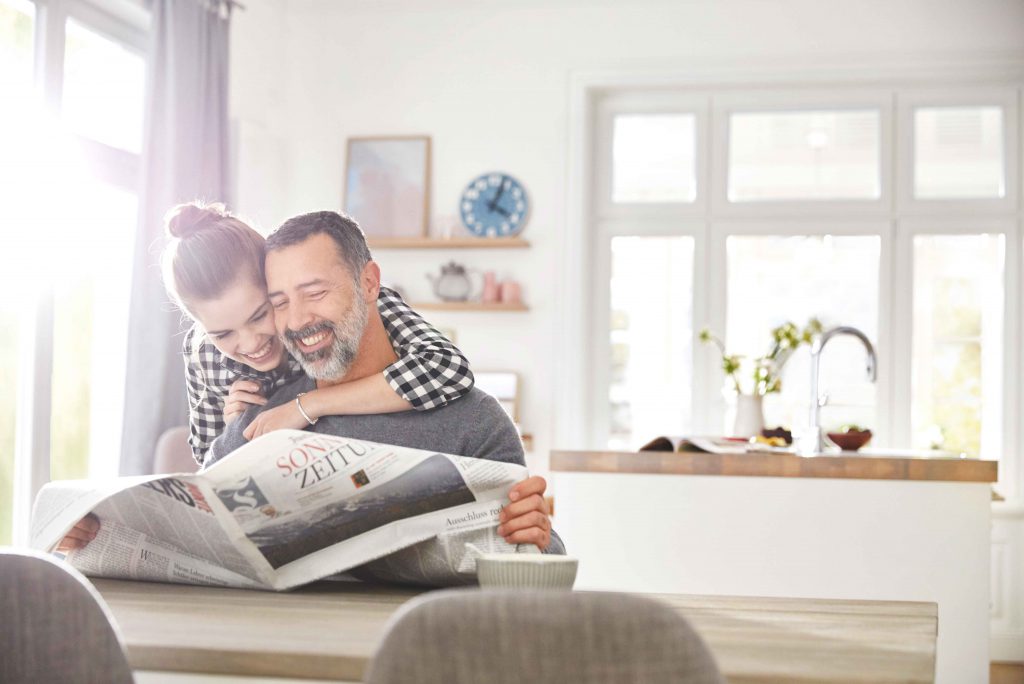 A girl hugs her father, who is sitting at a table reading a newspaper